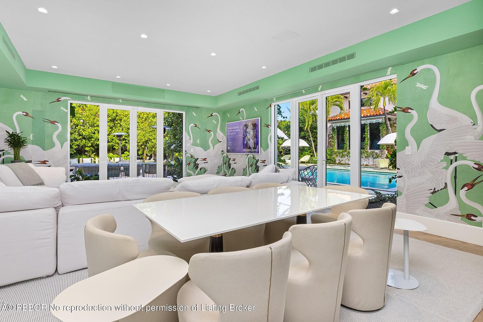 This bright and airy dining room features a large, glossy white table surrounded by modern, cream-colored upholstered chairs. The walls are adorned with a distinctive green wallpaper depicting large white cranes, creating a bold, nature-inspired aesthetic. Large glass doors provide a seamless transition to the outdoor pool area, filling the space with natural light and offering a serene view of the tropical landscaping.