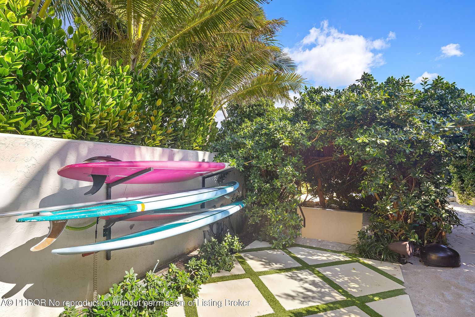 This outdoor space features a wall-mounted rack holding three surfboards, including a vibrant pink board and two blue-toned boards. The area is landscaped with lush green foliage and palm trees, set against a paved walkway with grass-filled geometric pavers. The scene captures a relaxed, coastal lifestyle aesthetic with bright natural sunlight.