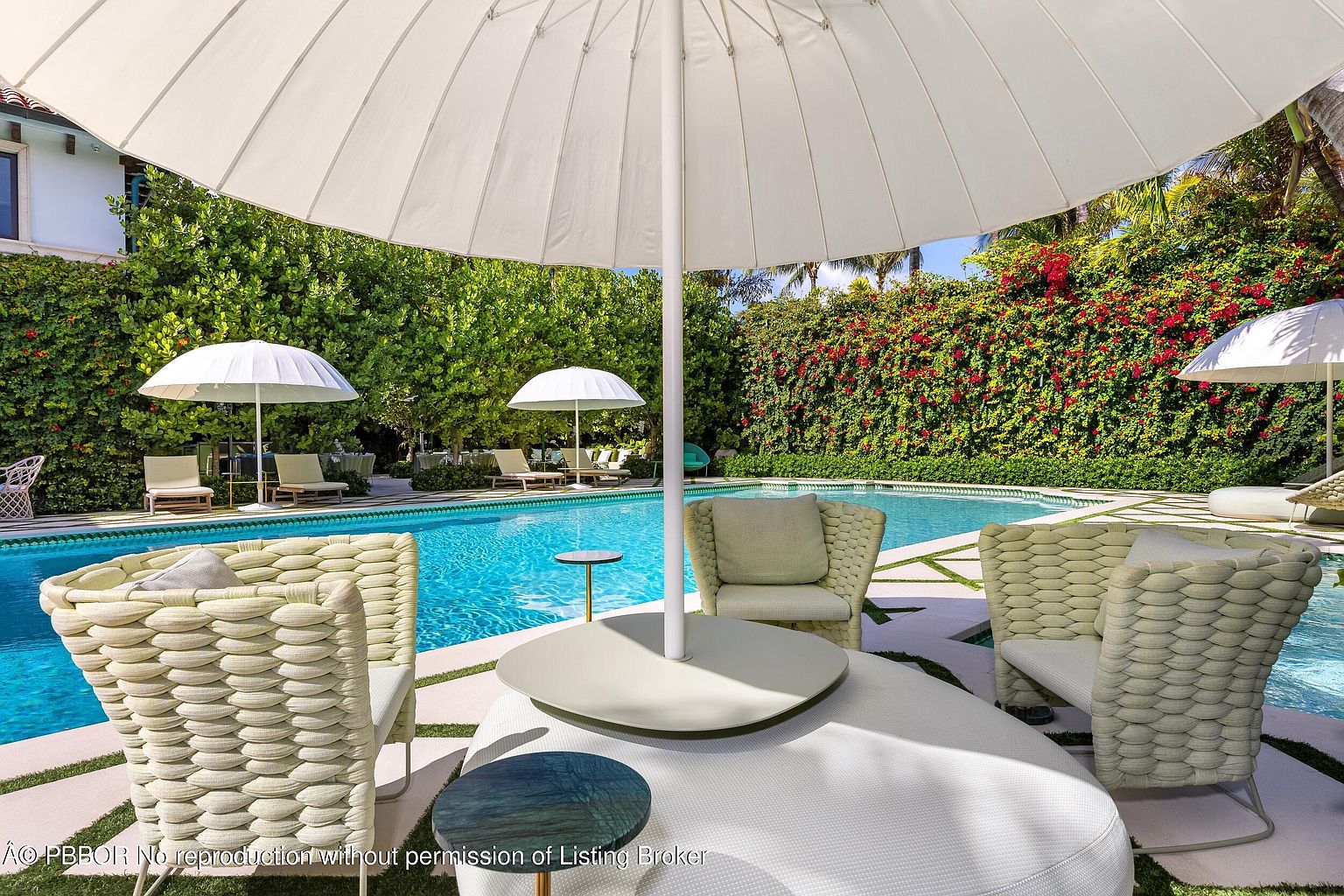 This luxurious outdoor pool area features a vibrant blue swimming pool surrounded by a paved deck with integrated greenery. The foreground showcases comfortable, woven-style patio furniture arranged under a large white umbrella, creating an inviting lounge space. Lush, dense hedges with blooming red flowers provide a private and serene backdrop to the resort-like setting.