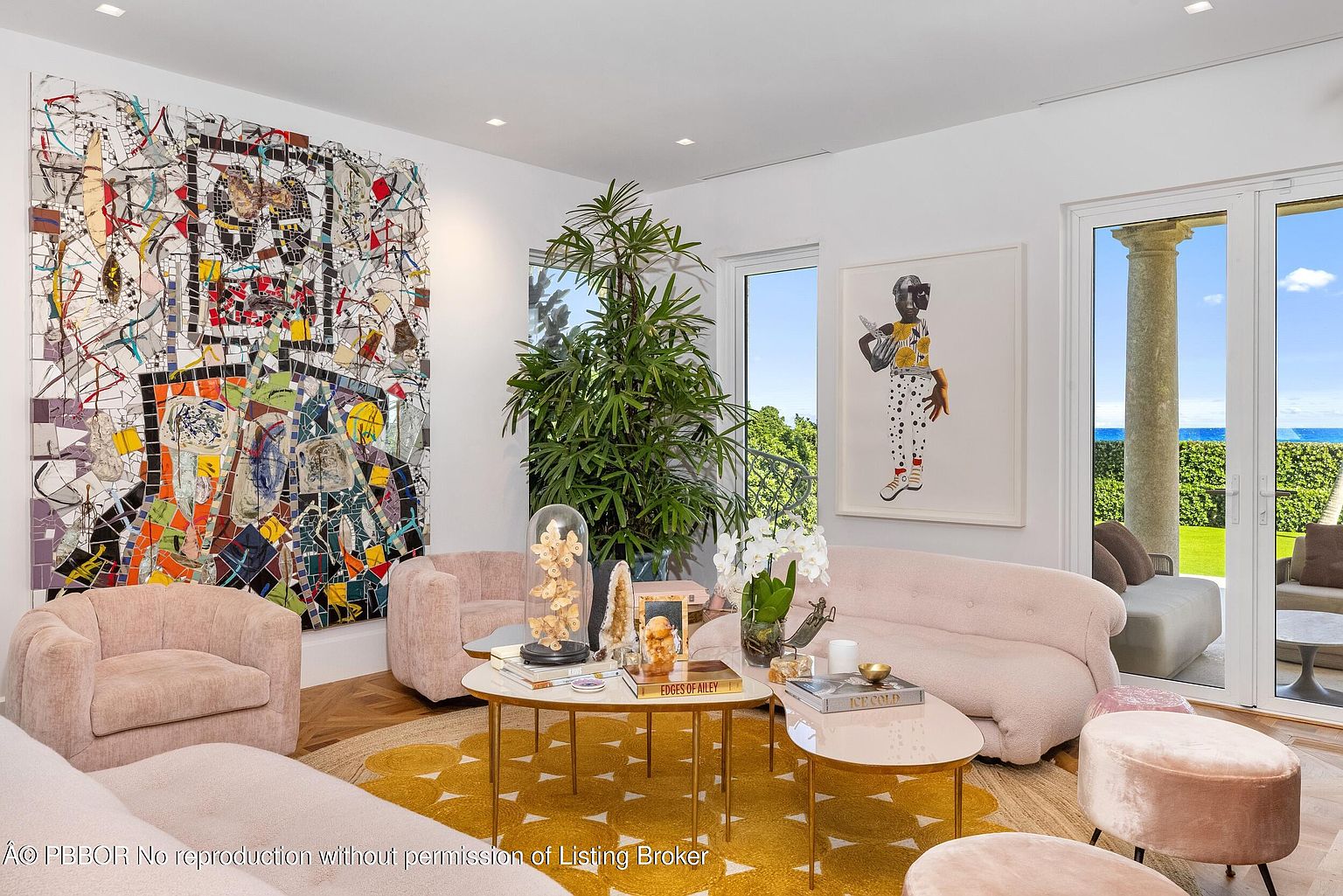 This sophisticated living room features a vibrant, large-scale mosaic art piece on the wall and a contemporary portrait painting, creating a gallery-like atmosphere. The space is furnished with plush, light-toned curved sofas and velvet ottomans arranged around a pair of gold-framed coffee tables atop a bold yellow patterned rug. Large glass doors provide a seamless transition to an outdoor patio with ocean views, while a tall indoor plant adds a touch of natural greenery to the bright, airy interior.