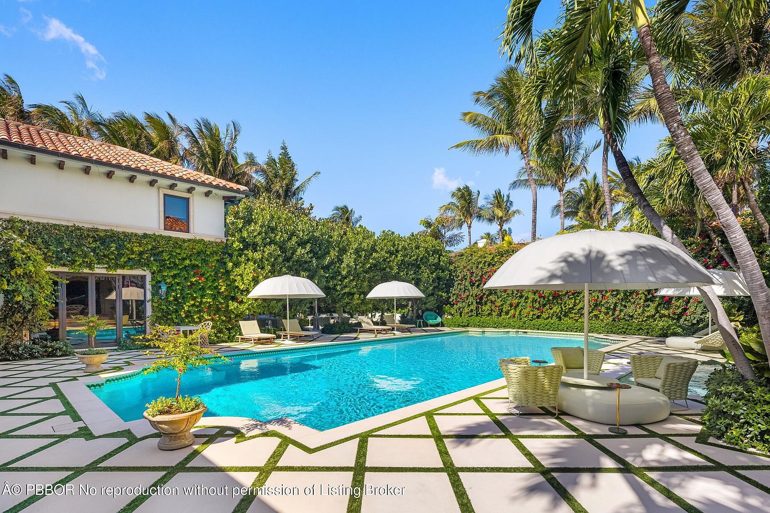This luxurious outdoor space features a large, rectangular swimming pool surrounded by a geometric stone patio with integrated grass grid patterns. The area is framed by lush, vine-covered walls and mature palm trees, creating a private, resort-like atmosphere. Multiple white umbrellas and lounge chairs are arranged around the pool, offering a serene setting for relaxation and entertainment.