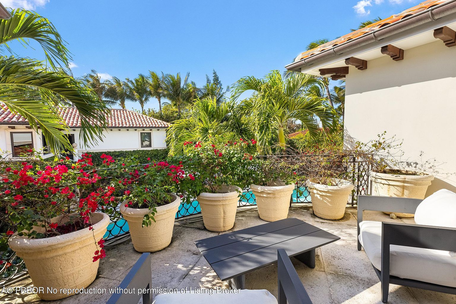 This elevated balcony offers a serene outdoor retreat, featuring stone flooring and a collection of large, light-colored planters filled with vibrant bougainvillea. A modern, dark-toned coffee table and comfortable lounge seating create a relaxing atmosphere, while a wrought-iron railing overlooks a lush garden and a glimpse of a swimming pool below. The scene is bathed in bright, natural sunlight, highlighting the Mediterranean-inspired architecture of the home.