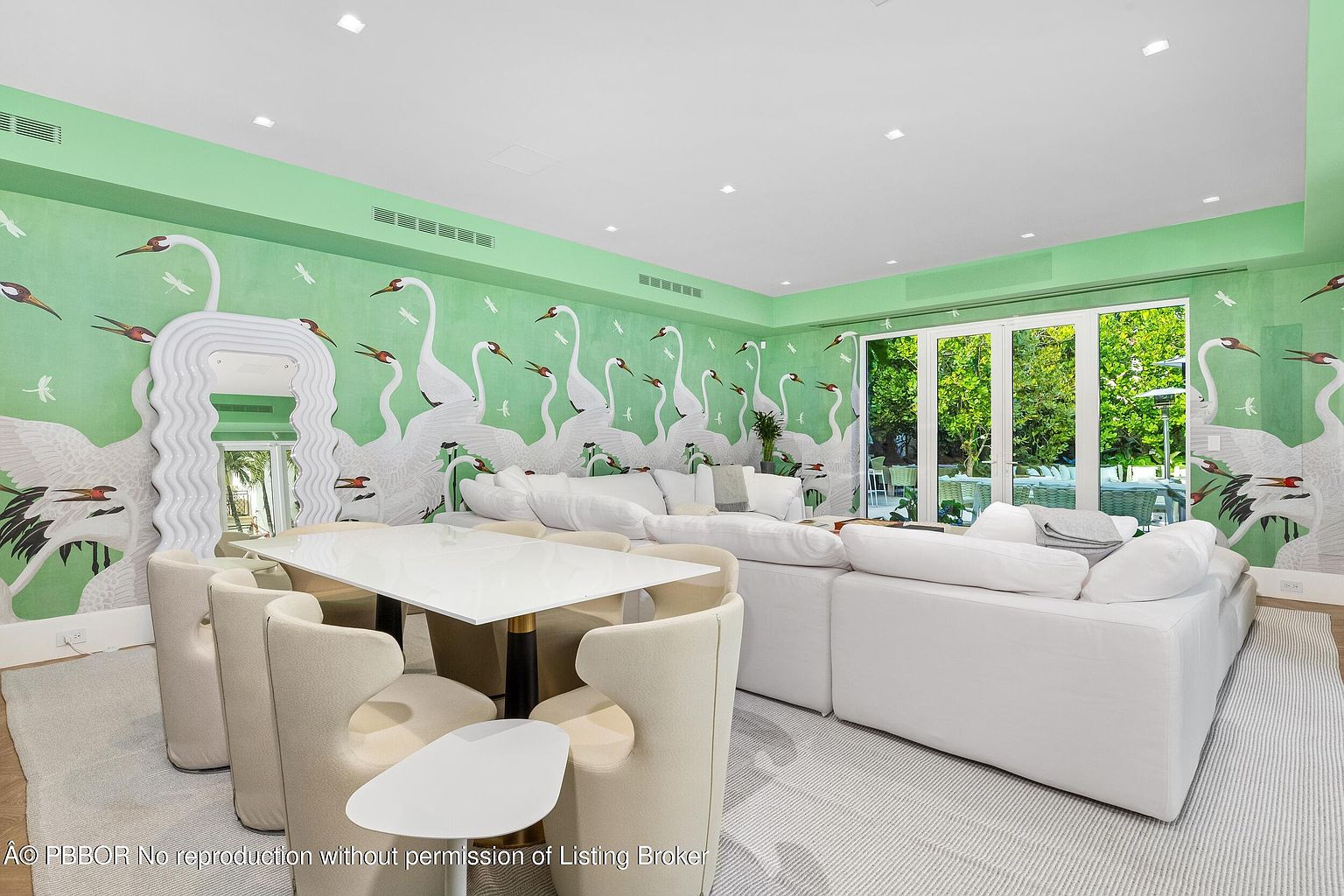 This vibrant living room features a bold, green wallpaper adorned with a large-scale crane motif, creating a whimsical and artistic atmosphere. The space is furnished with a plush, white sectional sofa and a modern dining set with cream-colored chairs, all set atop a textured light-colored rug. Large glass doors provide a seamless transition to the outdoor area, while a unique, wavy-framed mirror adds a contemporary sculptural element to the room.