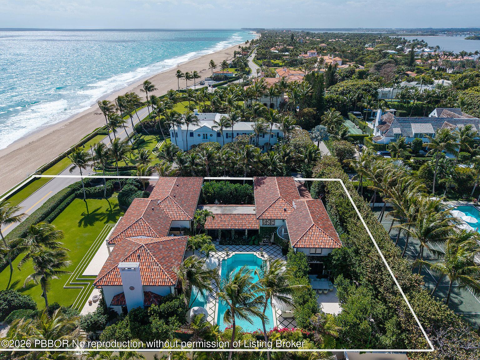 This high-angle aerial view captures a sprawling, U-shaped Mediterranean-style estate situated along a coastal road. The property features a central courtyard with a swimming pool, a red-tiled roof, and lush landscaping, all bordered by a white perimeter line. The expansive oceanfront setting provides a dramatic backdrop, showcasing the proximity to the beach and the surrounding upscale residential neighborhood.