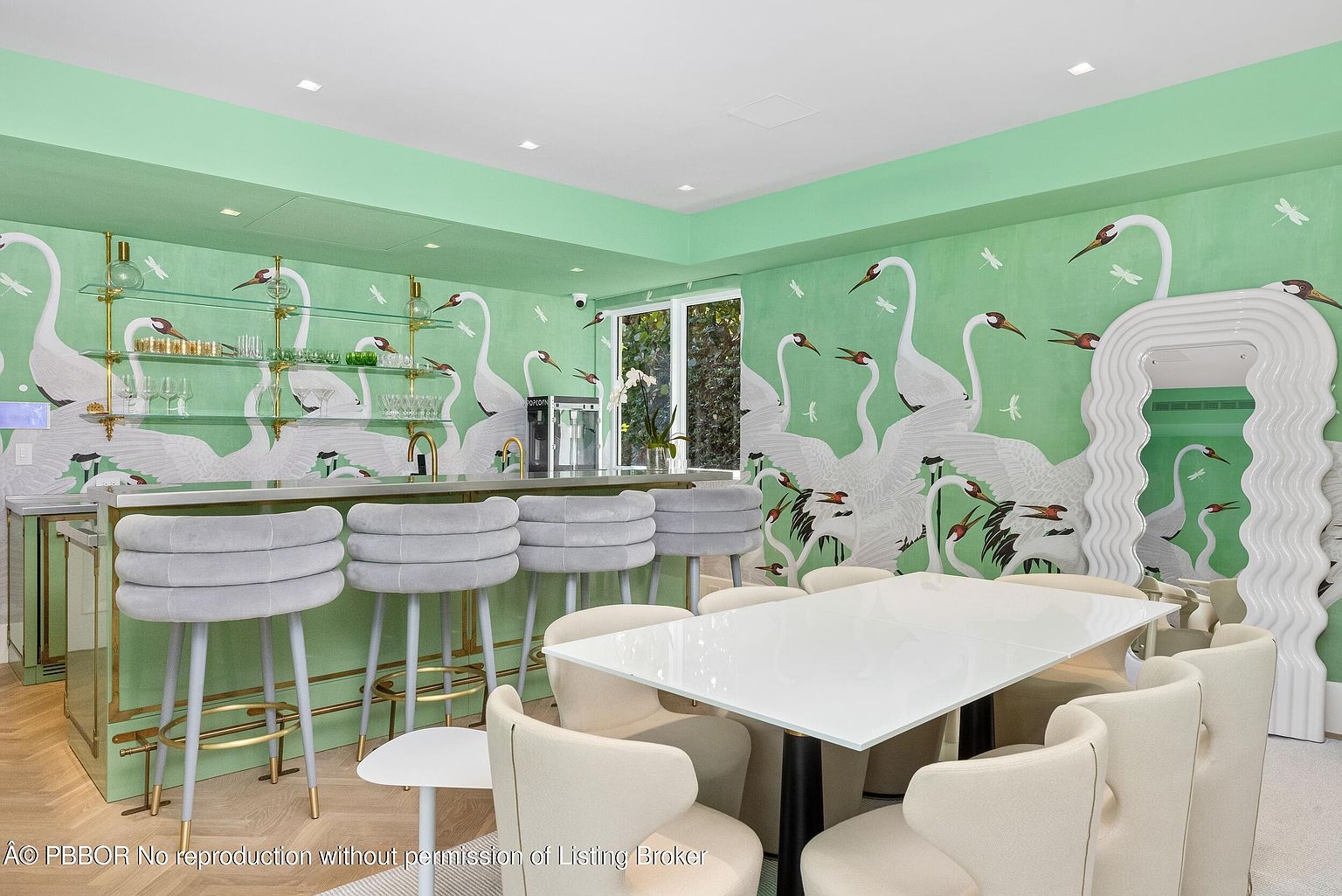 This vibrant dining and bar area features whimsical, crane-patterned wallpaper in shades of green and white, creating a bold and artistic atmosphere. The space includes a sleek bar with light-colored, upholstered stools and glass shelving, alongside a modern dining table surrounded by matching cream-colored chairs. A large, wavy-framed mirror adds a sculptural element to the room, while the light wood flooring provides a warm foundation for the eclectic decor.