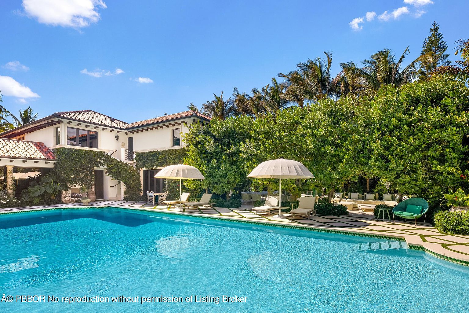 This luxurious outdoor space features a large, sparkling swimming pool as the centerpiece, surrounded by a geometric patterned stone patio. The area is furnished with multiple lounge chairs and umbrellas, set against a backdrop of lush green trees and a white, Mediterranean-style home covered in climbing vines. The scene captures a serene, resort-like atmosphere perfect for relaxation and outdoor entertaining.