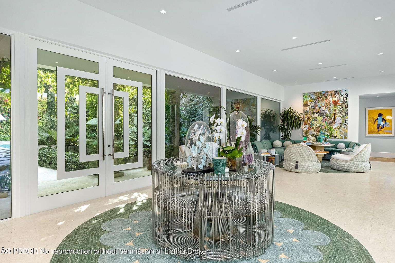 This bright and airy living space features a central circular metal table topped with decorative glass cloches and orchids, set upon a patterned teal rug. Large glass doors provide a seamless transition to the lush greenery outside, while the seating area in the background includes a plush green sofa and modern textured chairs. The room is characterized by its clean white walls, recessed lighting, and a sophisticated, contemporary aesthetic.