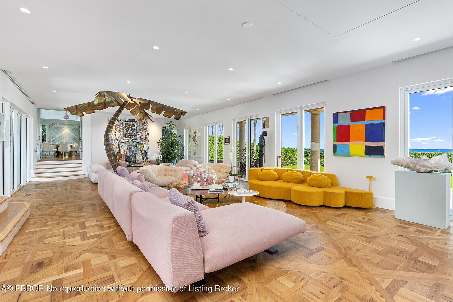 This expansive living room features a sophisticated open-concept layout with rich, patterned wood flooring and a bright, airy atmosphere. The space is furnished with a large, soft pink sectional, a vibrant yellow curved sofa, and a unique sculptural palm tree art piece, all set against a backdrop of large windows that provide abundant natural light and views of the outdoors. The room is decorated with colorful abstract wall art and a large crystal specimen on a pedestal, creating a luxurious and artistic living environment.