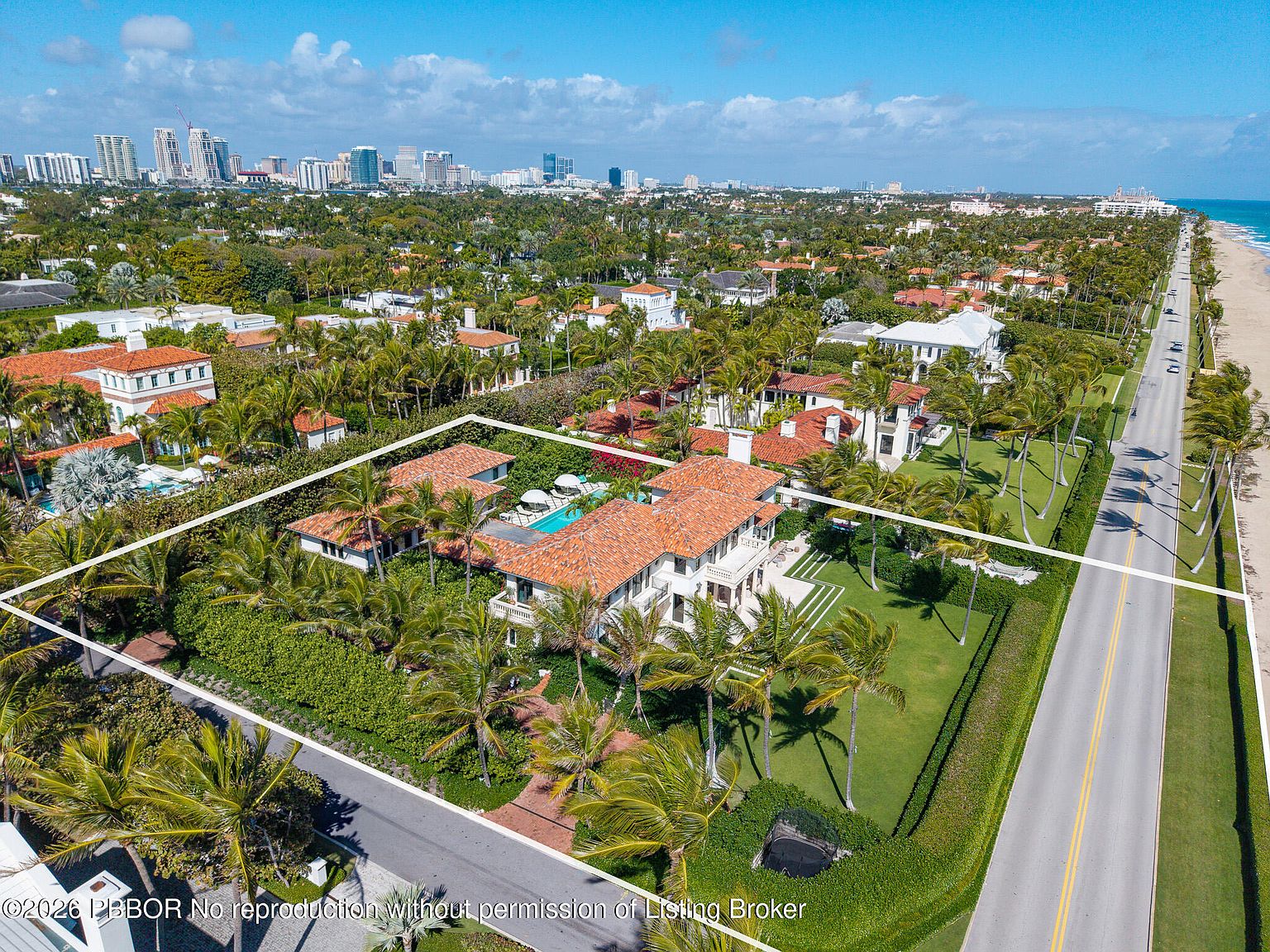 This high-angle aerial view captures a sprawling Mediterranean-style estate situated along a coastal road, bordered by a sandy beach and the ocean. The property features a terracotta-tiled roof, a central courtyard with a swimming pool, and lush tropical landscaping. The perspective highlights the home's prime location, showcasing the expansive grounds and the surrounding neighborhood with a distant city skyline on the horizon.