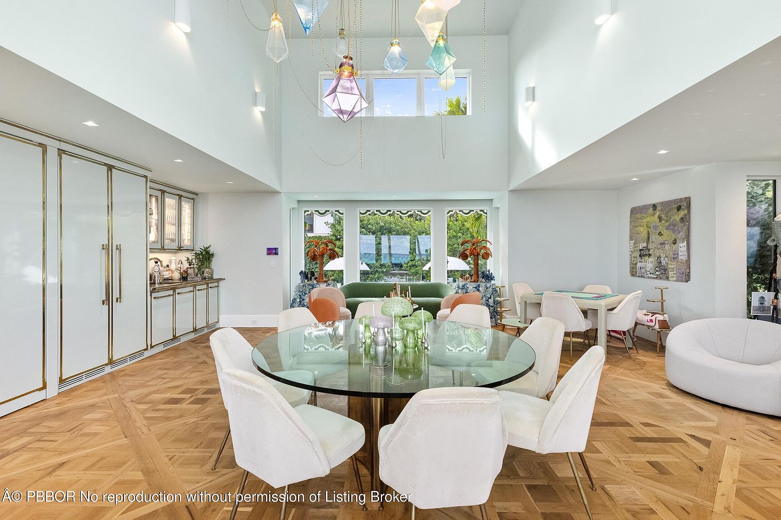 This expansive, high-ceilinged dining room features a round glass-top table surrounded by eight upholstered chairs, set upon a light-toned herringbone wood floor. A striking collection of colorful, geometric pendant lights hangs from the vaulted ceiling, while a wet bar with glass-front cabinetry is positioned along the left wall. Large windows at the rear provide a view of the lush greenery outside, creating a bright and airy atmosphere that flows into a secondary seating area.