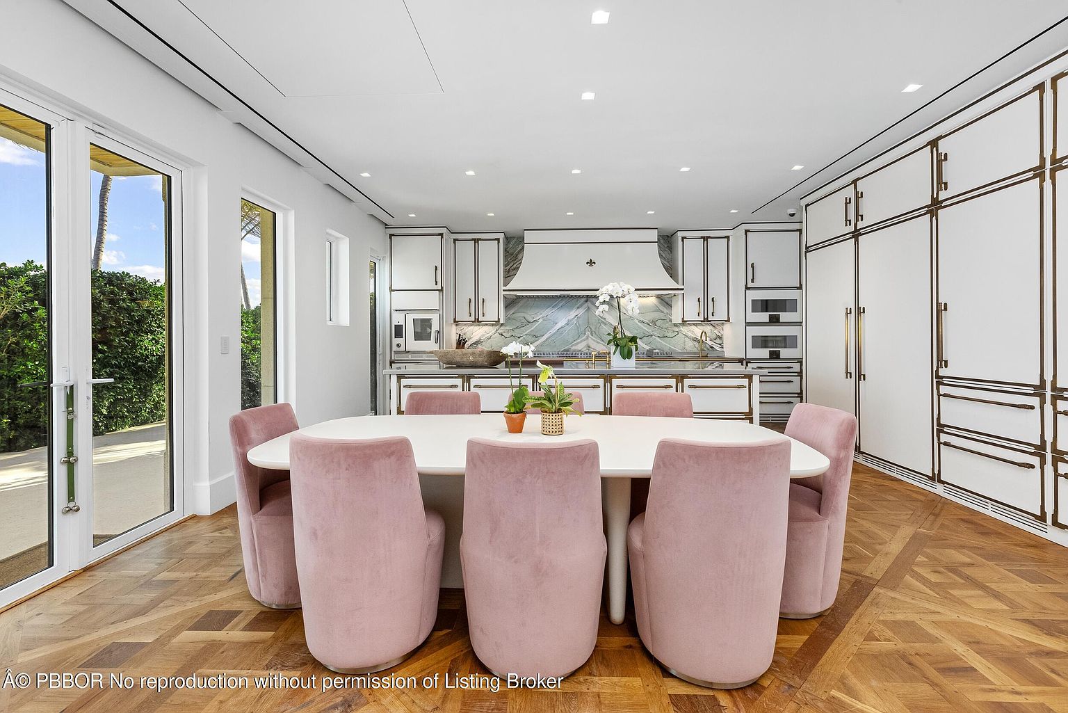 This bright, open-concept kitchen and dining area features a large white dining table surrounded by six soft pink upholstered chairs. The kitchen is equipped with white cabinetry accented by metallic trim, a stone-patterned backsplash, and a central island, all set against rich, patterned wood flooring. Large glass doors on the left provide a view of the outdoor greenery, creating a seamless transition between indoor and outdoor living spaces.