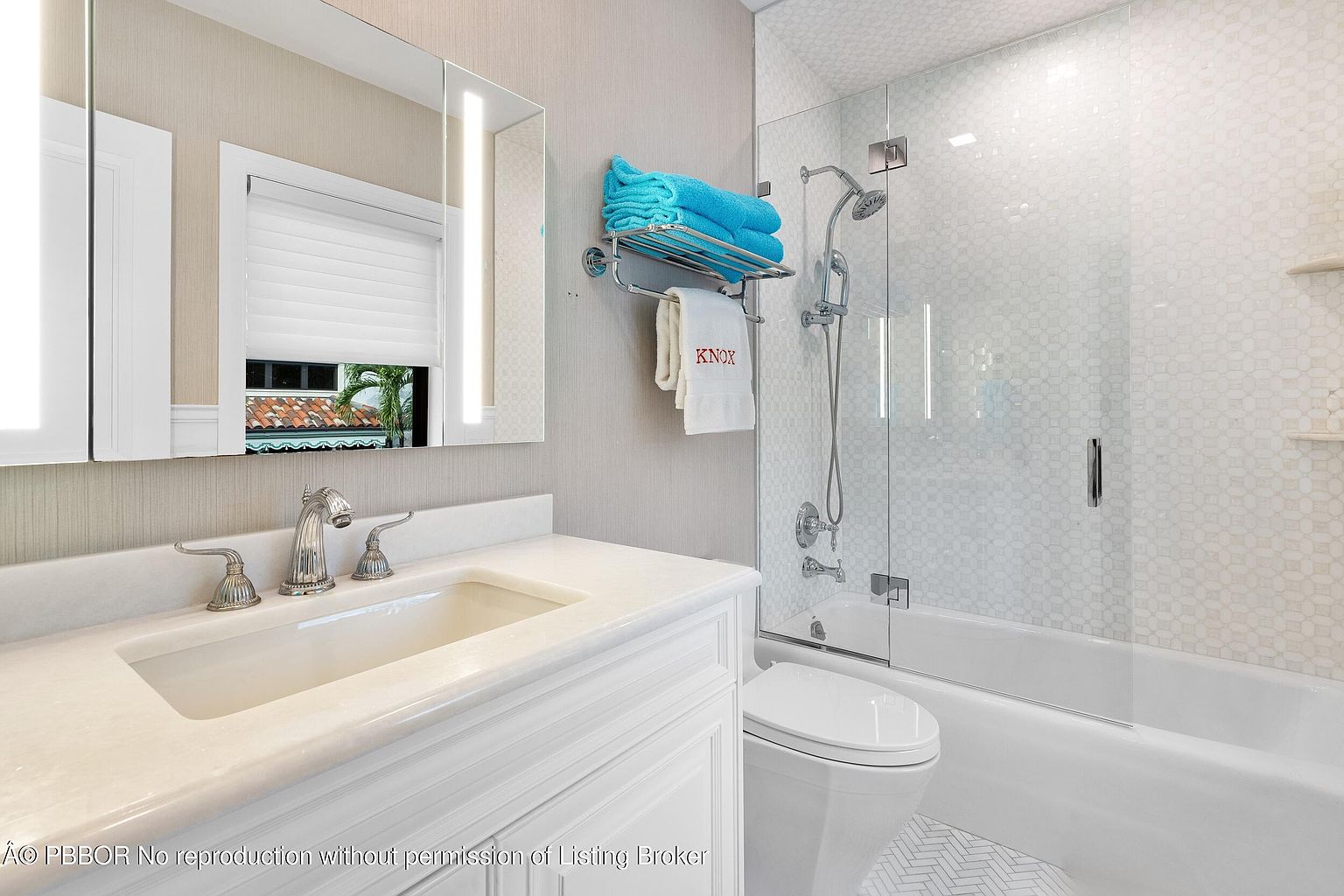 This bright, compact bathroom features a white vanity with a stone countertop and a polished chrome faucet. A glass-enclosed shower area with a patterned tile wall and a handheld showerhead sits adjacent to the toilet. The space is accented by a towel rack holding bright blue towels and a white towel embroidered with 'KNOX', creating a clean and functional aesthetic.