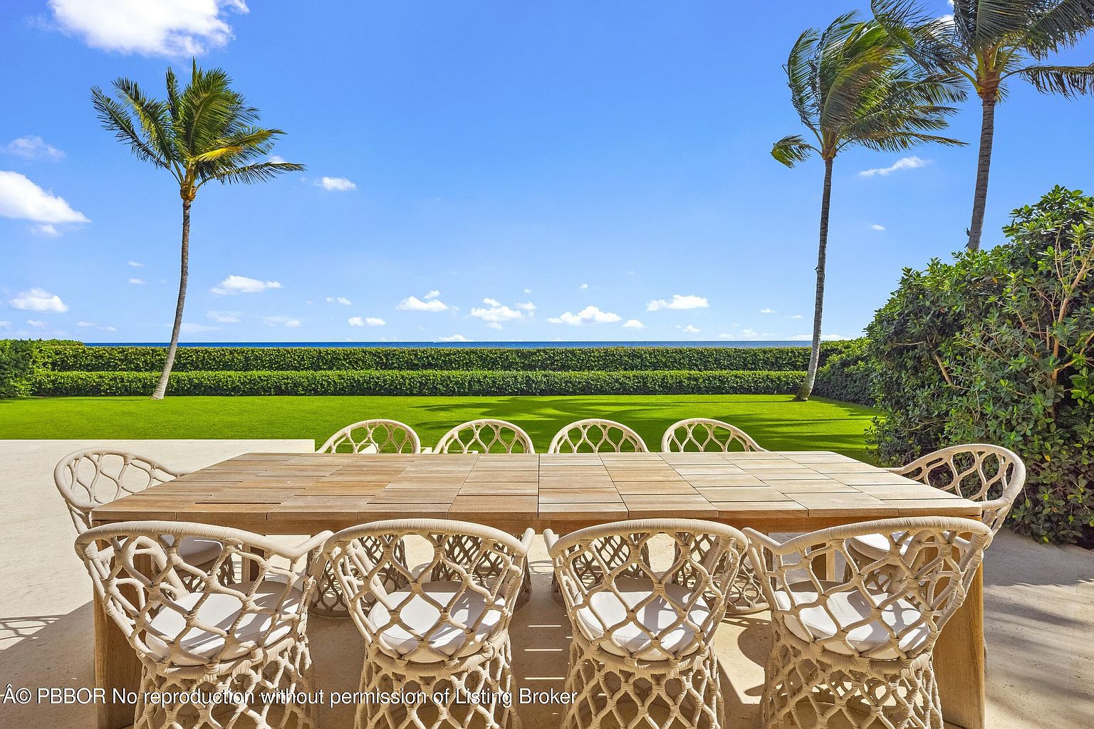 This outdoor dining area features a large, light-toned wooden table surrounded by woven rope chairs, set on a paved patio. The space overlooks a manicured green lawn bordered by a tall, neatly trimmed hedge, with two prominent palm trees framing a clear view of the ocean horizon. The bright, sunny atmosphere and open-air setting create an inviting and luxurious coastal living environment.
