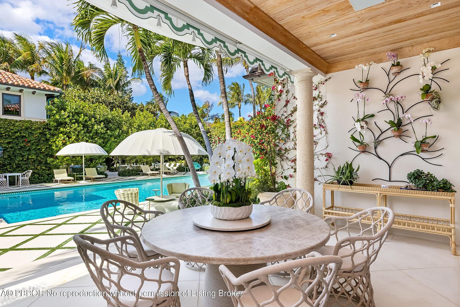 This elegant covered patio features a round stone-topped dining table surrounded by woven chairs, creating a sophisticated outdoor dining space. The area is accented by a decorative wall-mounted metal trellis adorned with potted orchids, while a light wood-paneled ceiling adds warmth to the structure. Beyond the patio, a pristine swimming pool is framed by lush tropical landscaping and white umbrellas, offering a serene and luxurious resort-like atmosphere.