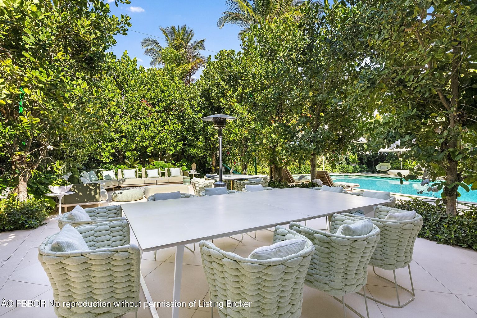 This expansive outdoor patio area features a large, modern dining table surrounded by woven-textured chairs, perfect for al fresco entertaining. The space is lushly landscaped with mature trees and greenery, creating a private oasis that transitions seamlessly into a sparkling swimming pool area. A tall outdoor heater stands near a secondary lounge seating arrangement, emphasizing the comfort and luxury of this resort-style backyard.