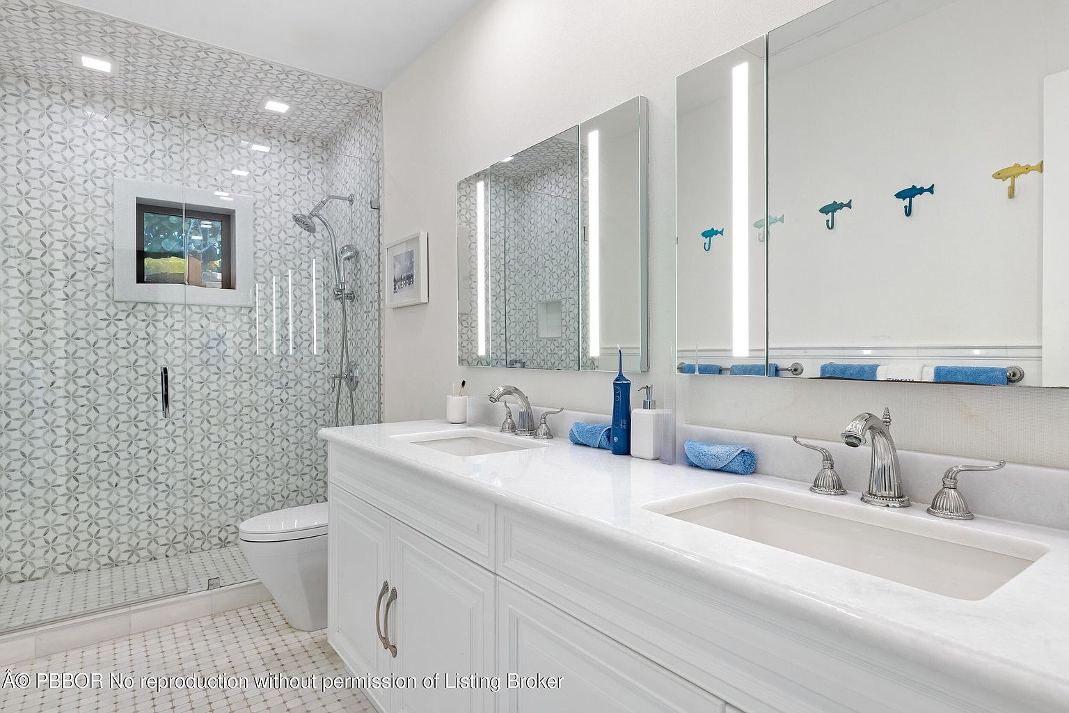 This bright, modern bathroom features a double vanity with a white countertop and dual sinks, complemented by large, illuminated mirrors. To the left, a walk-in shower is enclosed by a glass panel and finished with decorative patterned tile work. The space is clean and well-lit, with playful fish-shaped wall decor adding a touch of personality to the neutral color palette.