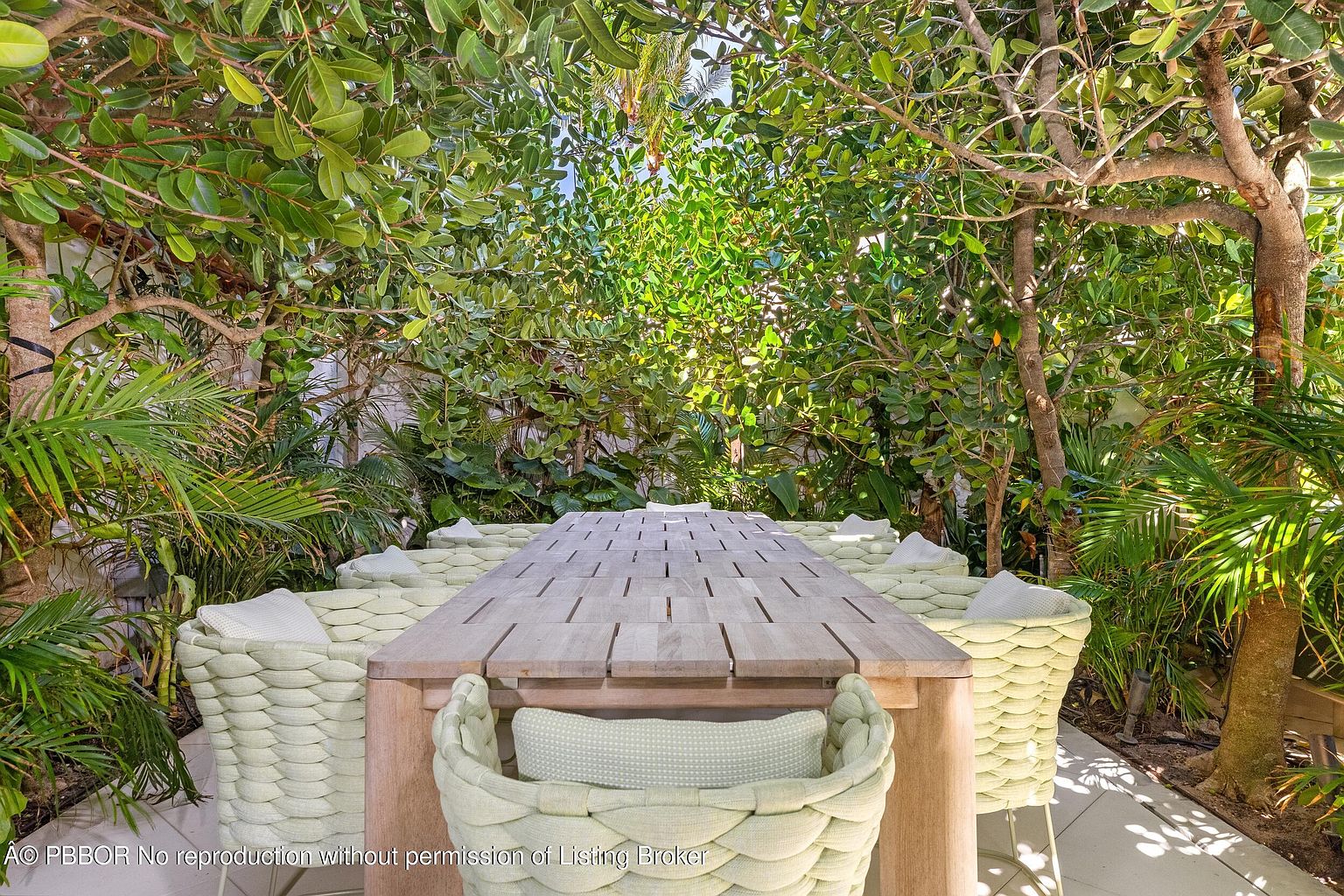 This outdoor dining area features a long, light-toned wooden table surrounded by woven-textured chairs with light-colored cushions. The space is nestled within a lush, dense garden setting, creating a private and serene atmosphere. The perspective is centered on the table, emphasizing the intimate and shaded nature of this al fresco dining spot.