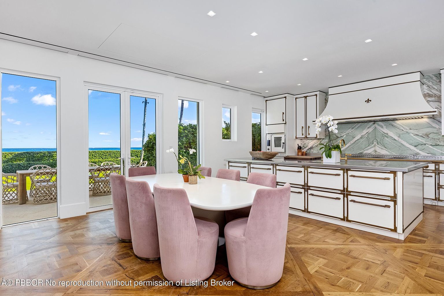 This bright, open-concept kitchen and dining area features a large central island with a stone countertop and a dining table surrounded by six soft pink upholstered chairs. The space is defined by elegant white cabinetry with gold-toned hardware, a striking green-veined stone backsplash, and intricate herringbone-patterned wood flooring. Large glass doors provide a seamless transition to an outdoor patio with ocean views, creating an airy and luxurious atmosphere.