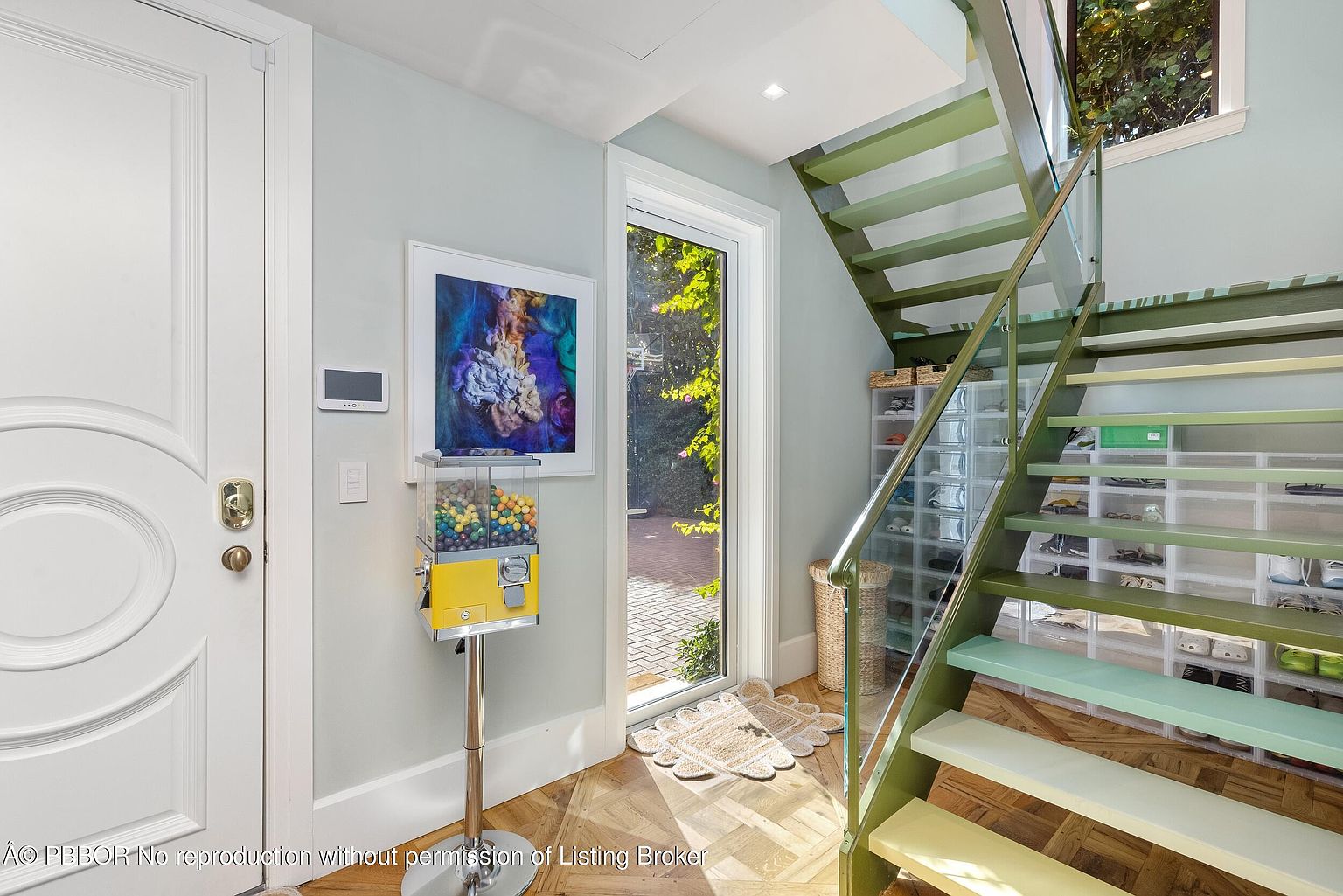 This bright entryway features a striking staircase with green-painted treads and a glass railing, creating a modern and airy aesthetic. Beneath the stairs, clear storage bins are neatly organized, while a vibrant piece of abstract art and a playful gumball machine add personality to the space. The light-colored walls and herringbone-patterned wood flooring lead toward a glass-paned door that opens to a lush outdoor view.