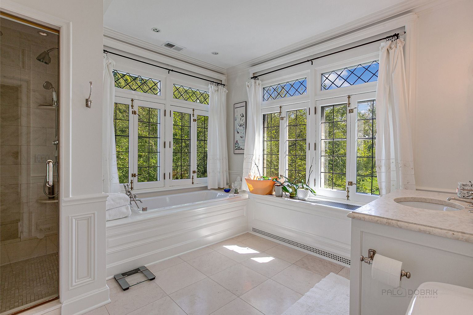 This is a bright and luxurious primary bathroom featuring a large soaking tub situated beneath two large windows with diamond-patterned panes, offering a view of lush greenery. The bathroom is decorated in a classic style with white wainscoting, marble countertops, and tile flooring. A glass-enclosed shower is visible to the left, and the overall impression is one of elegance and tranquility.