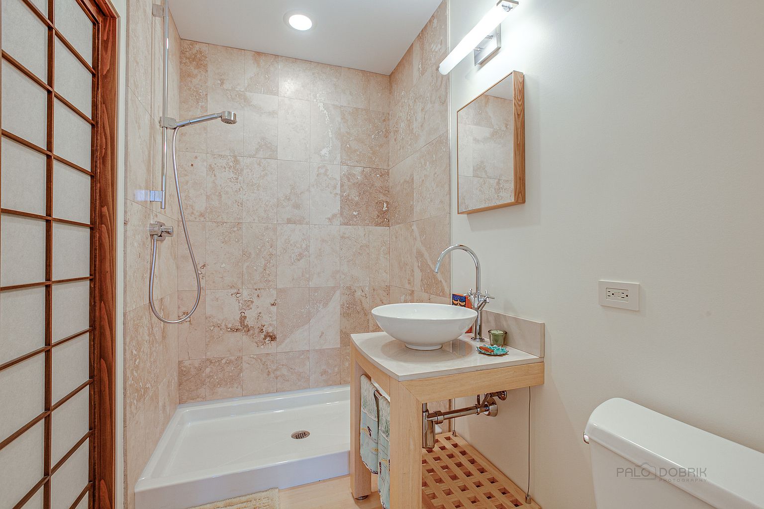 This is a well-lit bathroom featuring a walk-in shower with travertine tile, a modern vessel sink on a light wood vanity, and a framed mirror. The space is clean and functional, with a neutral color palette creating a calming atmosphere. The shower has a handheld shower head.