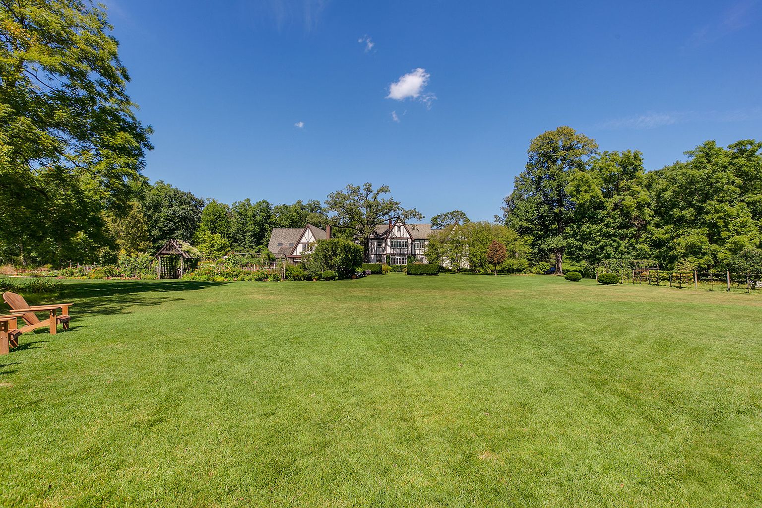 This image showcases a sprawling, well-manicured lawn leading up to a charming Tudor-style home. The property is surrounded by mature trees and lush landscaping, creating a serene and private setting. A wooden pergola and Adirondack chairs add to the inviting atmosphere of the yard.