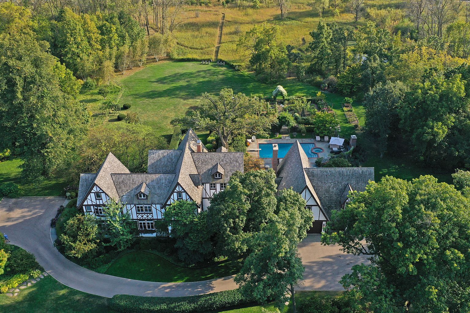 This aerial view showcases a grand Tudor-style home with multiple gabled roofs and half-timbered detailing. The property features a circular driveway, lush landscaping, a swimming pool with a spa, and extensive gardens. The surrounding mature trees and open fields create a private and serene estate setting.
