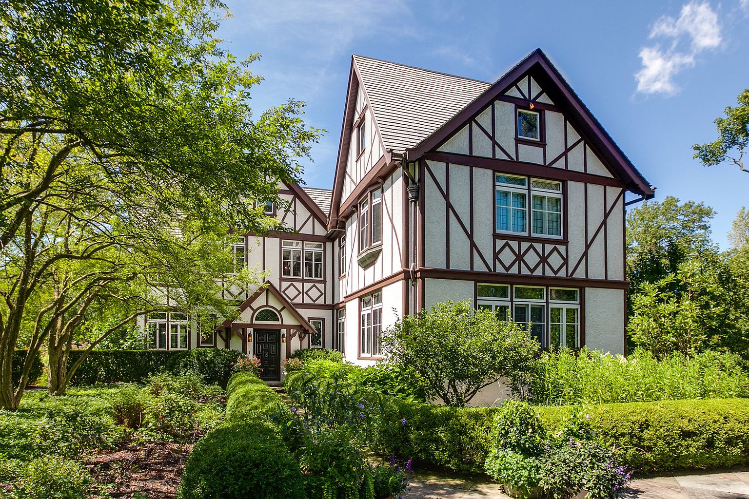 This is a front exterior view of a Tudor-style home with a well-manicured garden. The house features a white facade with dark brown trim, diamond-patterned detailing, and a steeply pitched roof. Lush greenery surrounds the property, creating a sense of privacy and charm.