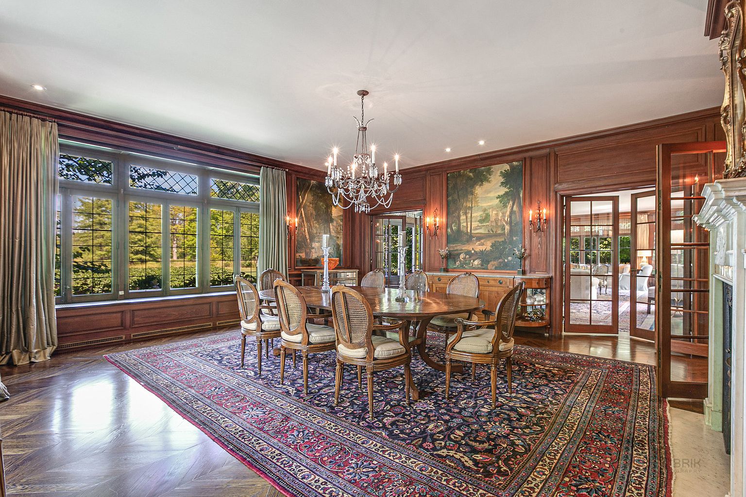 This is an elegant dining room featuring rich wood paneling, a large oval dining table with seating for eight, and a striking chandelier. A large window provides natural light and a view of the outdoors, while artwork adorns the walls. The room is furnished with a patterned rug that adds warmth and character to the space.