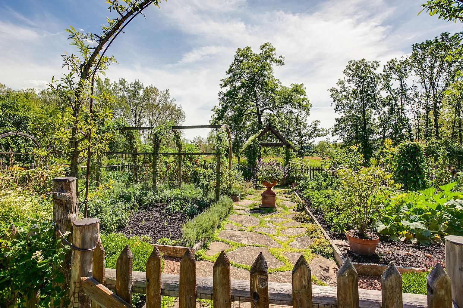 This image showcases a lush and well-maintained garden, likely part of a residential property. A stone pathway meanders through the garden, flanked by various plants, flowers, and wooden structures like trellises and a small arbor. A wooden picket fence in the foreground adds a charming, rustic touch, enhancing the garden's appeal and creating a sense of enclosure.