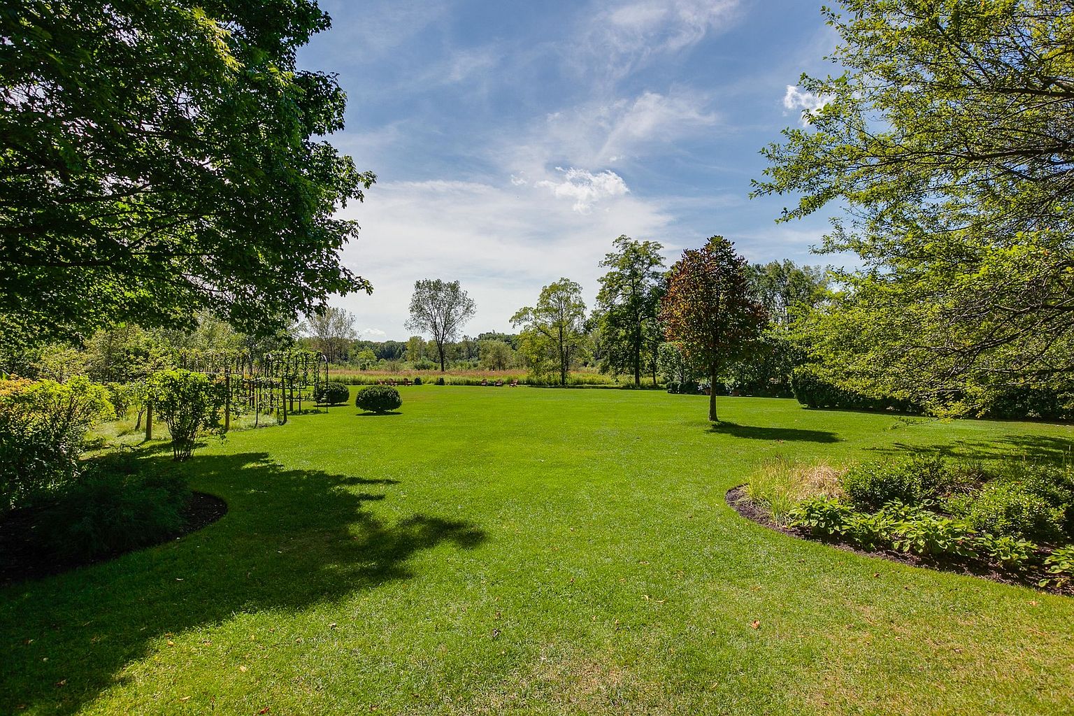 This image showcases a meticulously maintained yard or garden, featuring a lush green lawn that extends towards a line of trees in the background. The landscape includes various trees and bushes, adding depth and texture to the scene. The clear blue sky with scattered clouds enhances the overall appeal, creating a serene and inviting outdoor space.