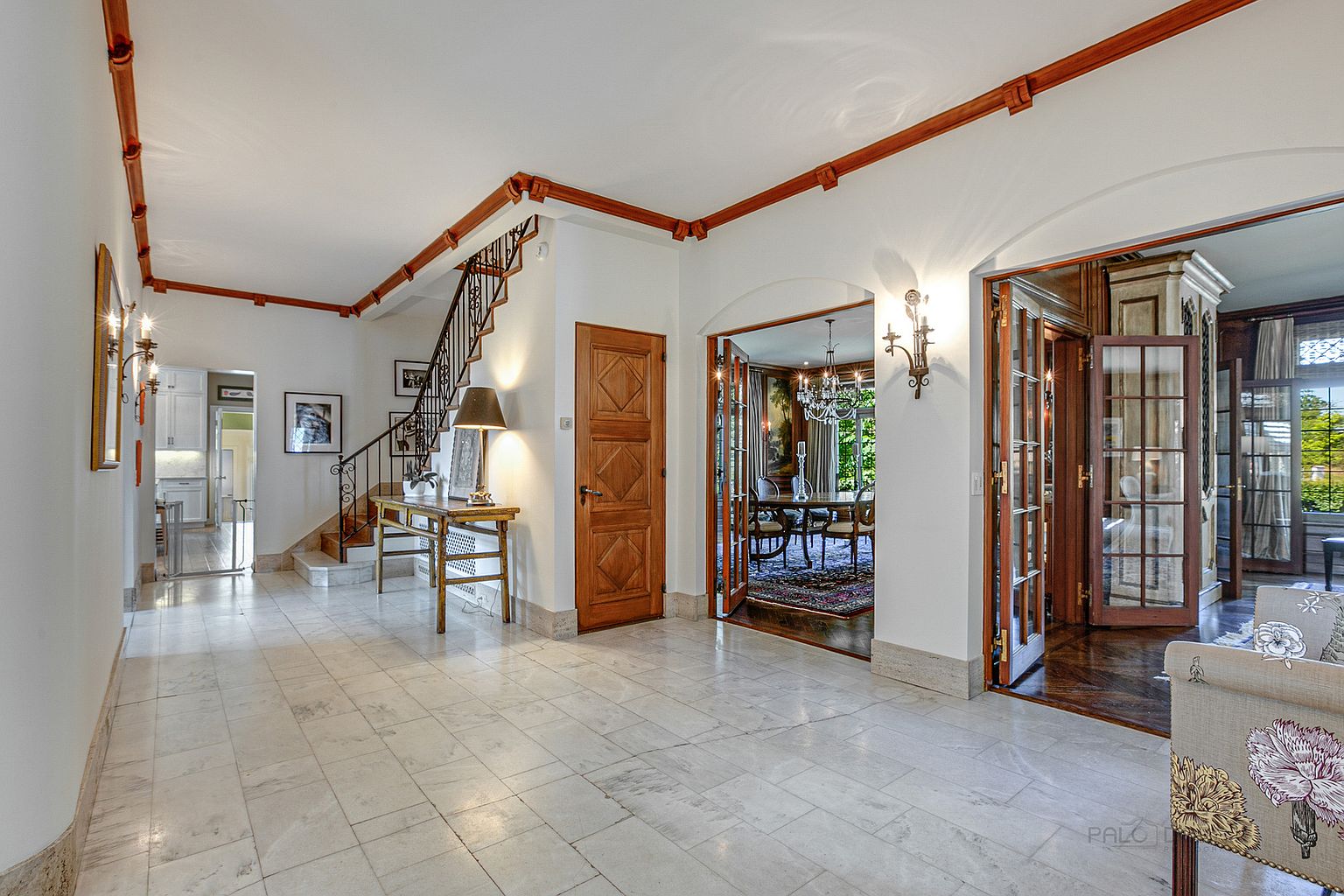 This interior shot showcases a grand hallway with marble flooring, a wooden staircase, and elegant archways leading to other rooms. The space is well-lit, highlighting the architectural details such as the wooden trim along the ceiling and the decorative wall sconces. The perspective gives a sense of depth and openness, emphasizing the home's sophisticated design.