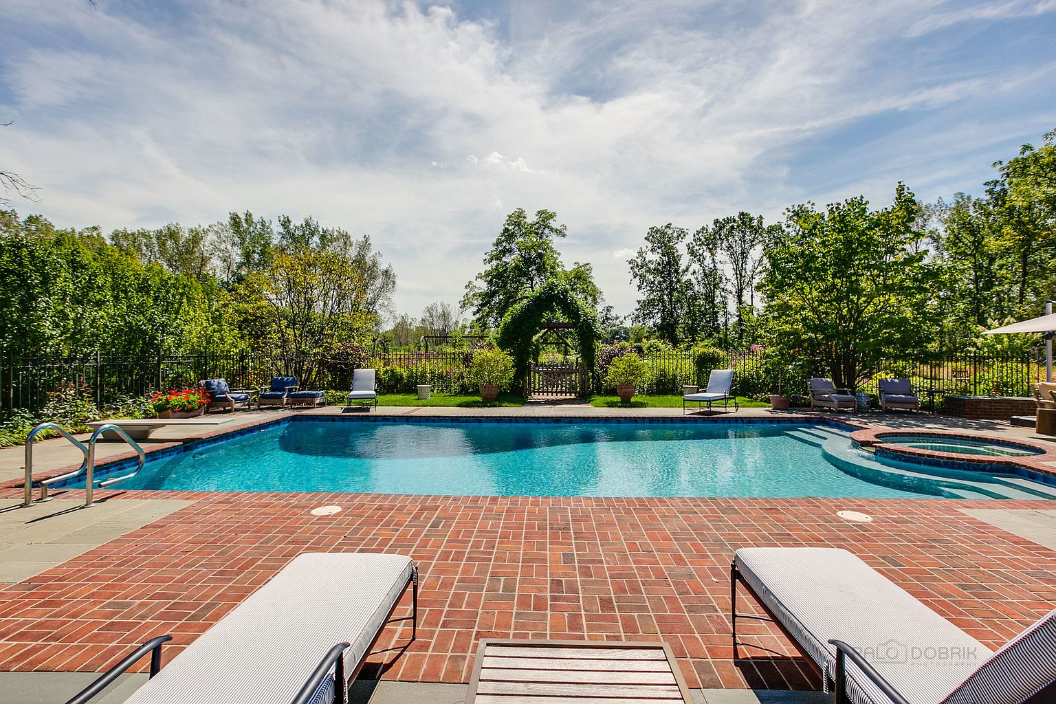 This image showcases a luxurious backyard pool and spa area. The pool is surrounded by a brick patio with lounge chairs, and a charming garden gate with an archway is visible in the background. The scene evokes a sense of relaxation and upscale outdoor living.