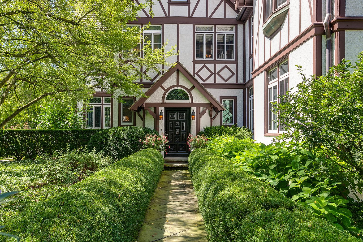 This image showcases the grand entryway of a Tudor-style home, featuring a meticulously manicured garden path leading to a dark, ornate front door. The house is characterized by its white stucco facade, dark wood trim, and diamond-patterned detailing. Lush greenery and mature trees frame the scene, creating an inviting and elegant first impression.
