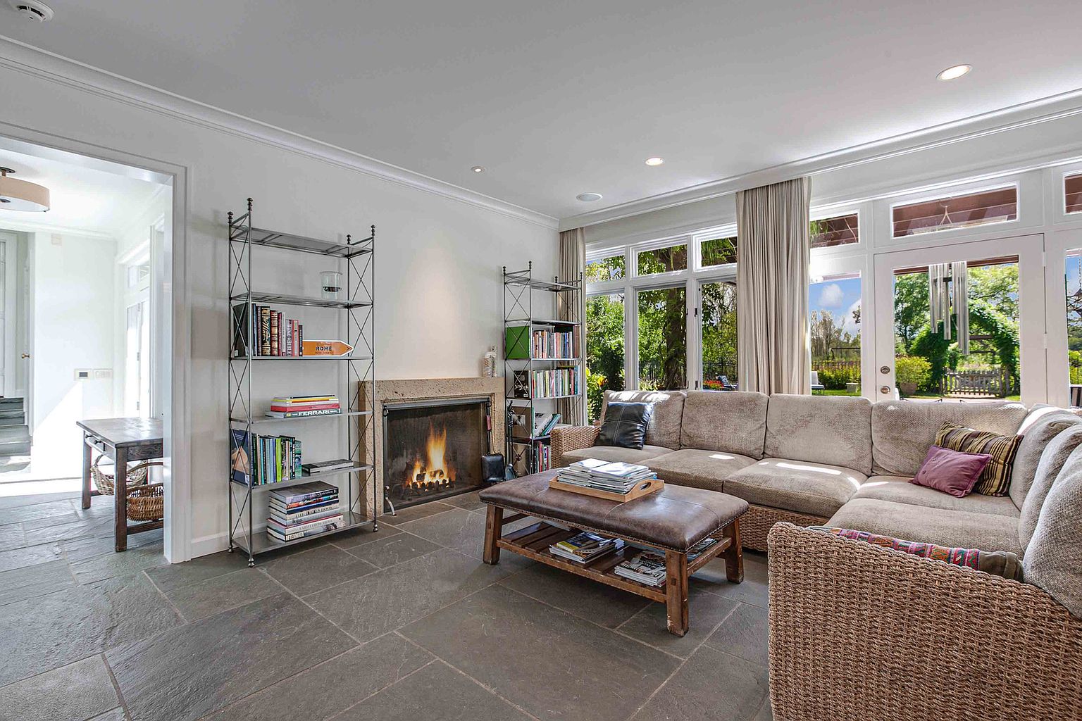 This is a cozy living room featuring a stone fireplace, built-in bookshelves, and a large sectional sofa. The room has a neutral color palette with natural light streaming in through large windows overlooking a garden. The flooring is stone tile, and the overall impression is one of comfort and relaxation.