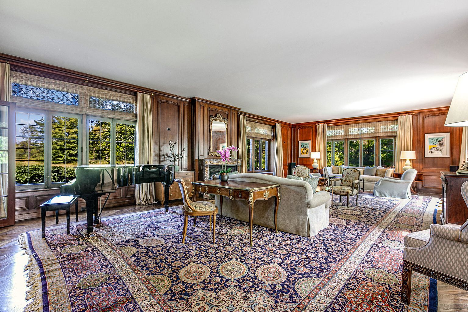 This is a luxurious living room featuring rich wood paneling, large windows offering natural light, and an ornate area rug that anchors the space. A grand piano sits near the windows, while comfortable seating arrangements are centered around a fireplace. The room exudes a classic and elegant ambiance, perfect for entertaining or relaxing.