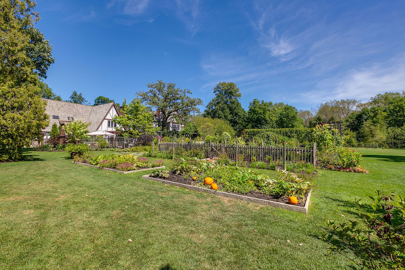 This image showcases a well-maintained garden with raised beds, lush greenery, and a variety of plants and pumpkins. A charming wooden fence encloses the garden area, providing a sense of privacy and definition. In the background, a glimpse of a house adds to the overall appeal of the property, suggesting a peaceful and inviting outdoor space.