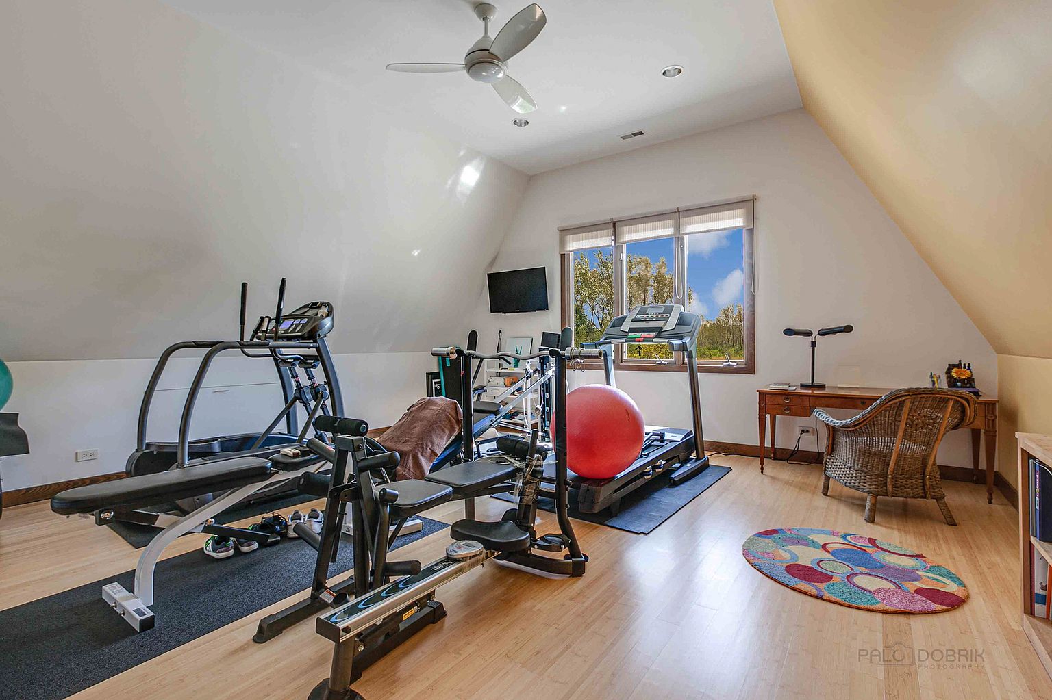This is an interior shot of a home gym featuring various exercise machines including treadmills, a weight bench, and a rowing machine. The room has hardwood floors, a ceiling fan, and a window providing natural light. A desk and chair are positioned near the window, suggesting a multi-functional space.