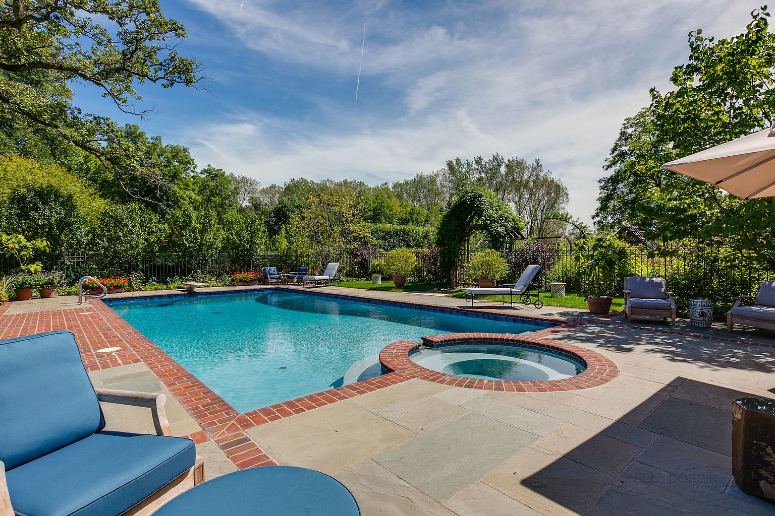 This image showcases a luxurious backyard pool and spa area, perfect for relaxation and entertainment. The pool features a classic rectangular design with a brick border, complemented by a circular spa. The surrounding patio is paved with large stone tiles, and lush greenery provides privacy and a serene atmosphere, enhancing the property's appeal.