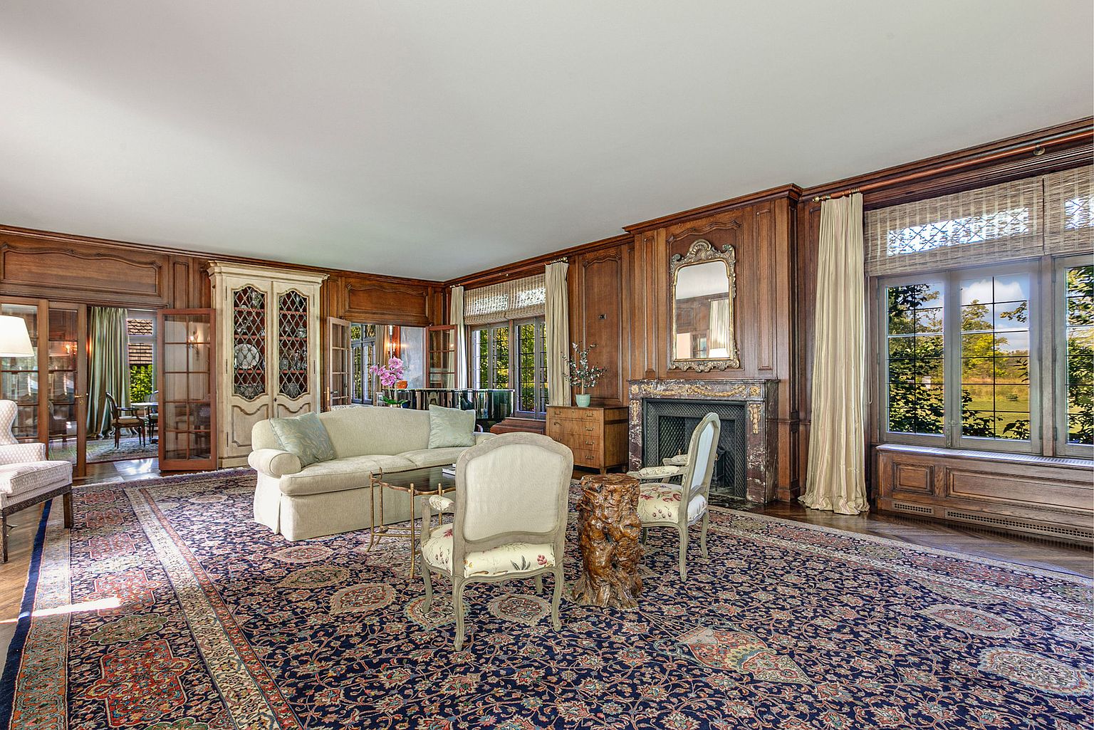 This is a spacious living room featuring rich wood paneling, a large oriental rug, and elegant furnishings. The room includes a fireplace with a decorative mirror above, a comfortable sofa, and two chairs arranged around a small table. Natural light streams in through the windows, creating a warm and inviting atmosphere.