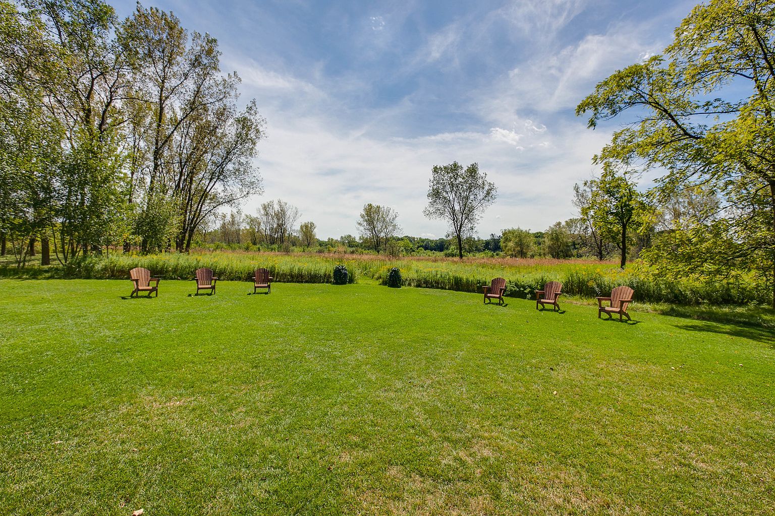 This image showcases a well-maintained yard or garden area, featuring a lush green lawn and several wooden Adirondack chairs arranged in a semi-circle. The setting is serene, with mature trees bordering the space and a natural meadow visible in the background, creating a peaceful and inviting outdoor space. The perspective is a wide shot, capturing the expanse of the yard and its integration with the surrounding landscape.