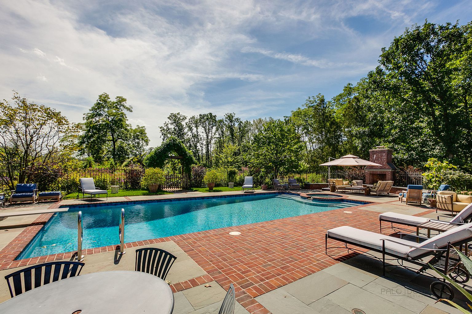 This image showcases a luxurious outdoor pool area, complete with a rectangular pool, brick patio, and various lounge chairs and seating arrangements. A brick fireplace and umbrella provide additional comfort and style. The surrounding landscape features lush greenery and trees, creating a private and serene backyard oasis.