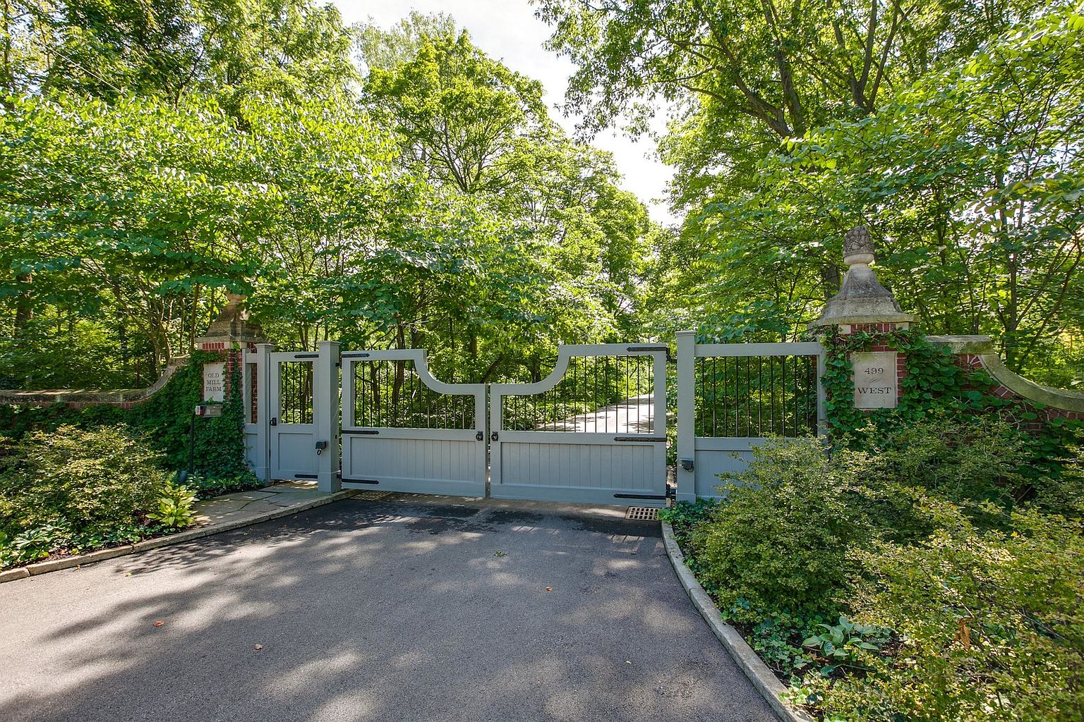 The image showcases an elegant gated entryway to a property, featuring a light gray wooden gate flanked by brick pillars adorned with ivy. The driveway leads through the gate and into a lush, green landscape. The overall impression is one of privacy and sophistication, suggesting a well-maintained and desirable estate.