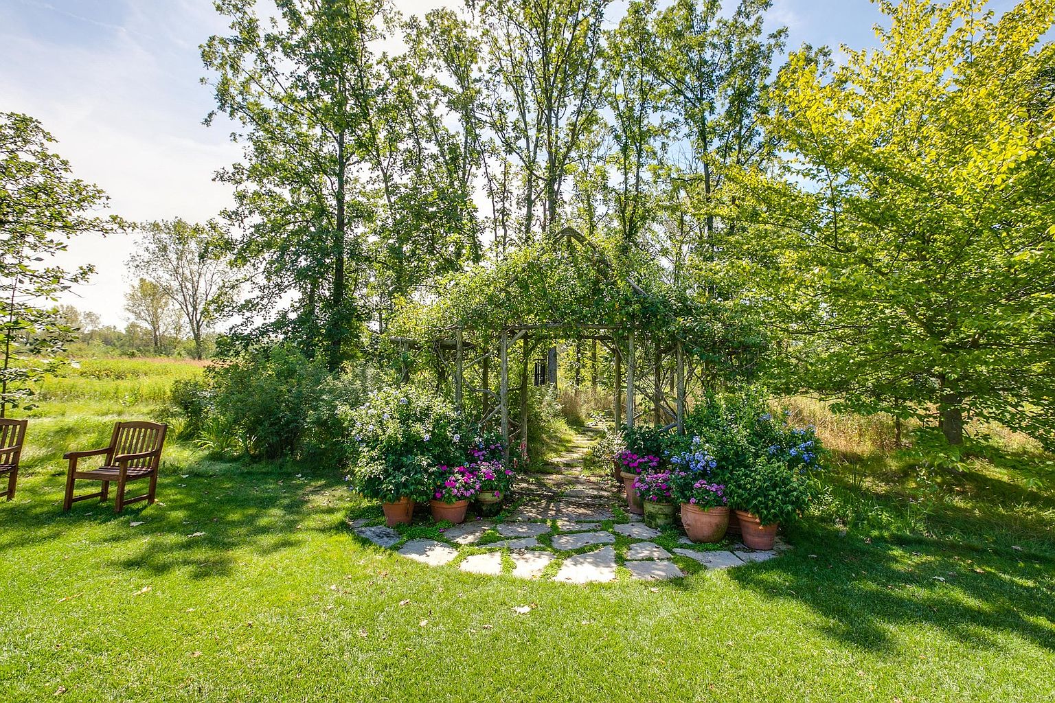This image showcases a beautifully landscaped garden with a charming wooden pergola covered in climbing plants. Potted flowers add pops of color, and a stone pathway leads through the structure. A wooden bench sits on the lush green lawn, creating a serene and inviting outdoor space.