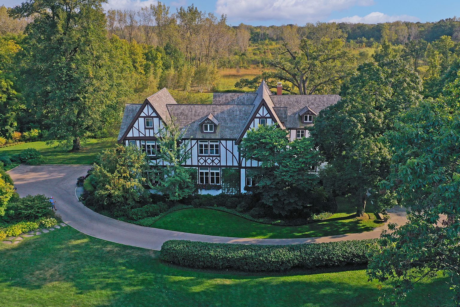 This aerial view showcases a grand Tudor-style home nestled among lush greenery. The property features a circular driveway, manicured lawns, and mature trees, creating a private and serene setting. The architectural details of the home, including its steeply pitched roofs and decorative timber framing, are prominently displayed.