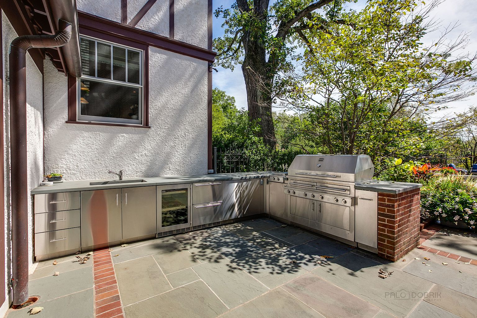 This image showcases an outdoor kitchen area on a patio. The kitchen features stainless steel cabinets, a built-in grill, a sink, and a beverage refrigerator, all set against the backdrop of a well-maintained yard. The patio is paved with stone tiles and accented with brick edging, creating an inviting space for outdoor entertaining.