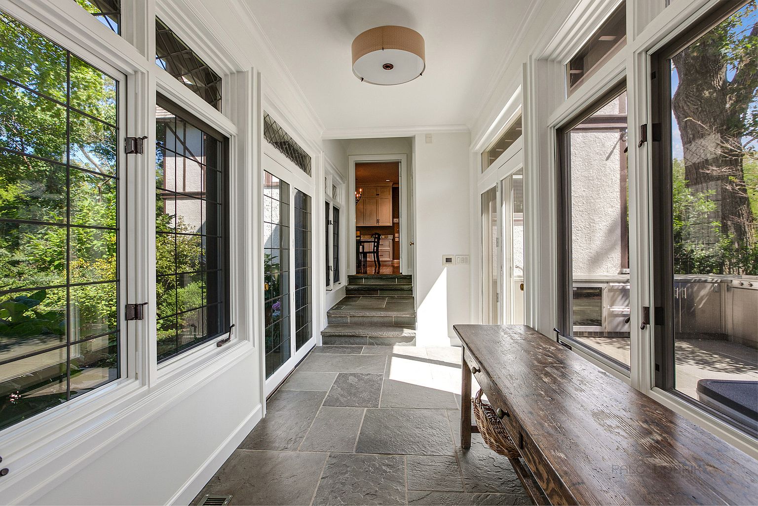 This interior shot showcases a bright sunroom or hallway with large windows offering views of the exterior greenery. The space features a stone tile floor, white trim, and a rustic wooden console table. A doorway leads to another room, accessible via a short set of stairs, adding depth and interest to the composition.