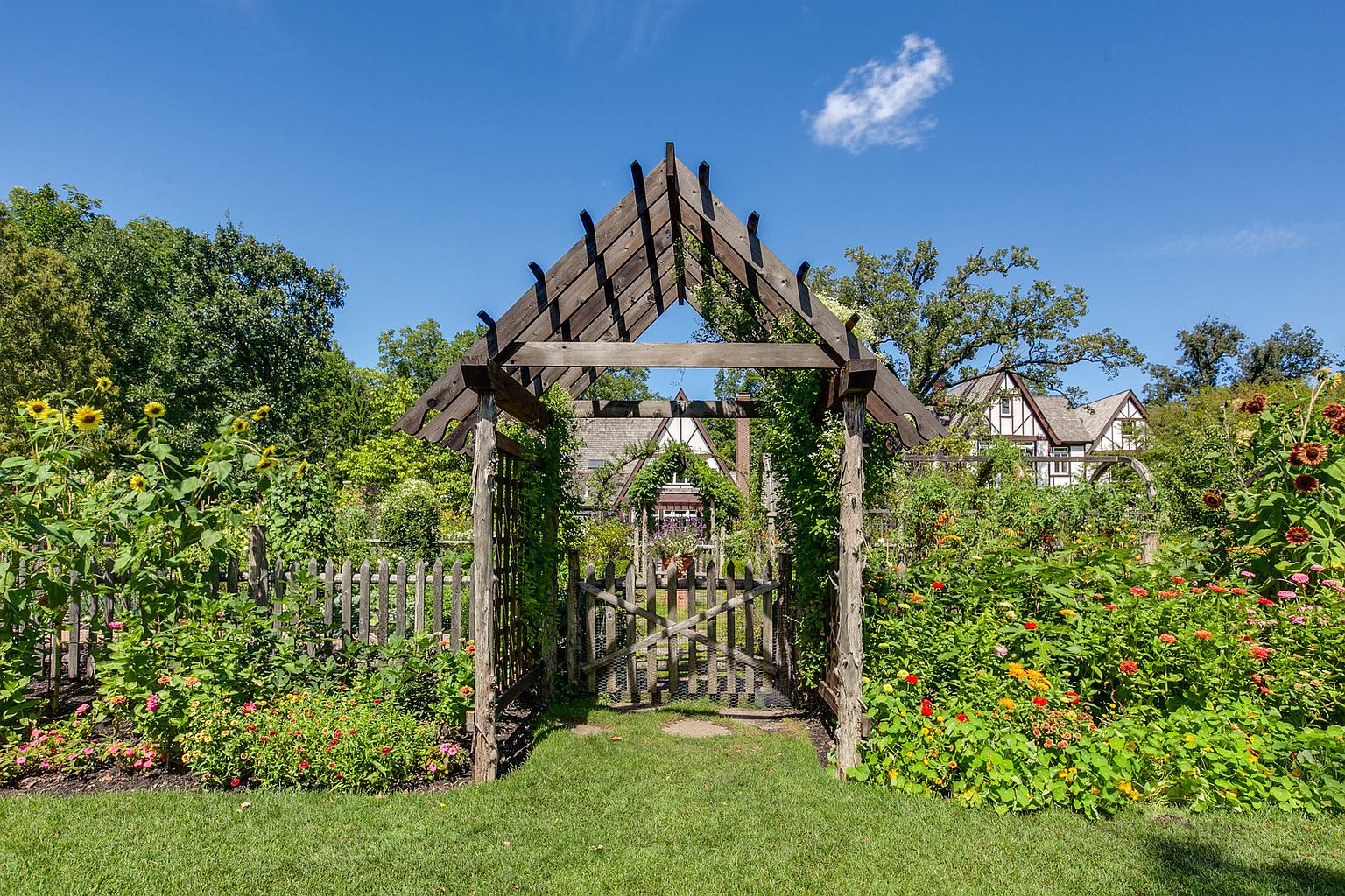 This image showcases a charming garden with a wooden pergola and gate, leading to a glimpse of a Tudor-style house in the background. The garden is lush with various flowers and greenery, creating a vibrant and inviting atmosphere. The perspective is from the lawn, looking through the gate, emphasizing the depth and beauty of the garden.
