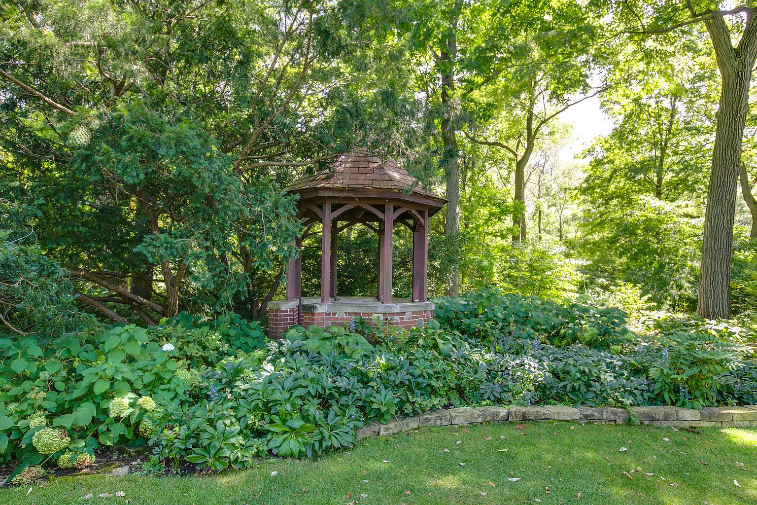 This image showcases a beautifully landscaped garden featuring a charming gazebo as a focal point. Lush greenery, including various plants and trees, surrounds the gazebo, creating a serene and inviting atmosphere. A well-maintained lawn and stone edging add to the garden's appeal, suggesting a meticulously cared-for outdoor space.