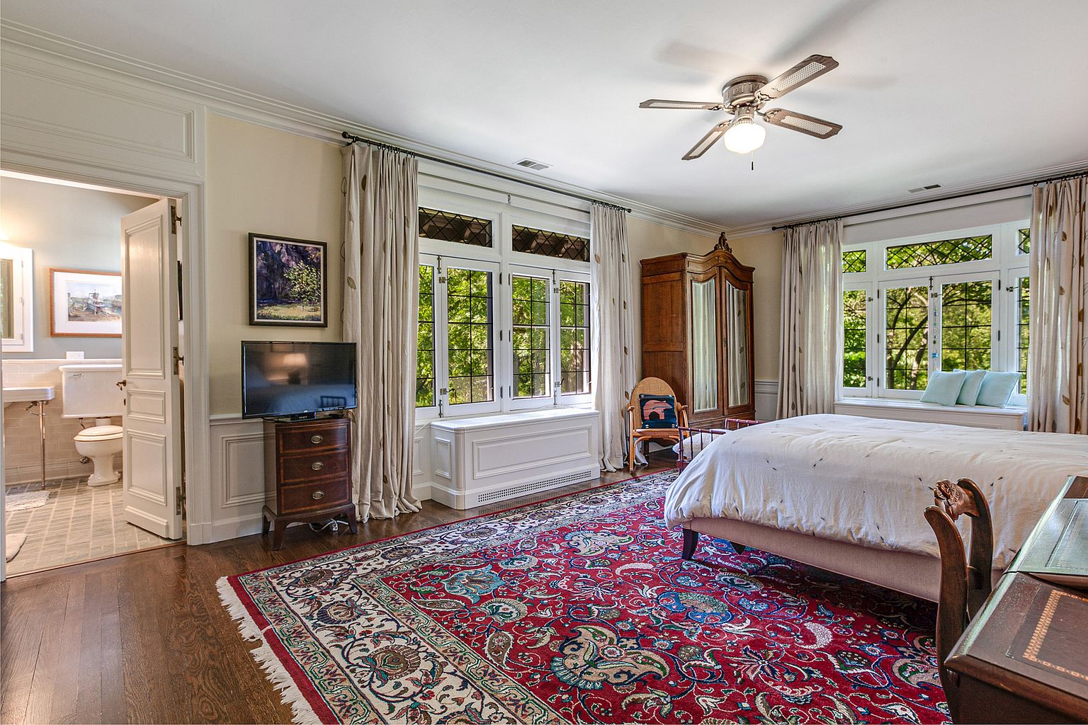 This is a primary bedroom featuring a large red patterned rug, a bed with white linens, and two windows with window seats. There is a television on a wooden dresser, a wardrobe, and a door leading to a bathroom. The room has a traditional style with detailed trim and neutral wall colors.